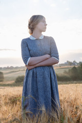 young girl in blue dress with collected hair poses, looks away in field of ears at sunrise. Foggy...