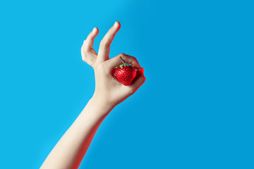 collage of bright juicy strawberries in hand on blue background. fingers show a sign okay