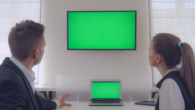 Business people in office. Back view employee tell about new project pointing on display with green screen on the wall. Businesswoman listening presentation attentively. Back view man and woman