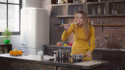 Happy adult woman cooking dinner tasting sauce in the contemporary kitchen. Lady standing near cooker with boiling saucepan. Smiling caucasian lady in flat.