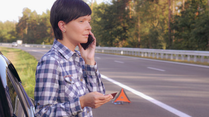 Young woman standing on the driveway near broken car holding card and mobile calling local garage or mechanic. Caucasian young female talking by smartphone on the roadside.