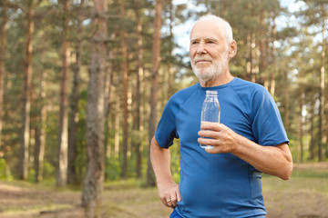 Portrait of handsome tired European senior elderly male in t-shirt holding glass bottle, enjoying...