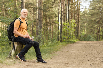 Obraz premium Summertime, hiking, active lifestyle and age concept. Energetic Caucasian man on retirement spending summer day outdoors in wild nature, traveling by foot, having rest on stump with bottle of water