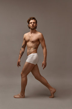 Full length shot of fit attractive tattooed man with muscular perfect body walking isolated wearing white boxer shorts. Handsome bearded guy demonstrating his muscles, having confident look
