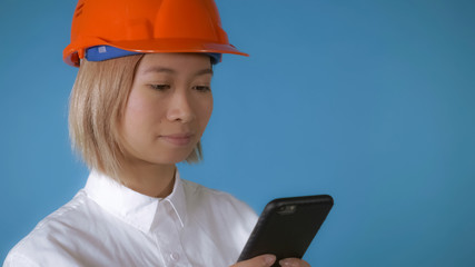 close up face young asian female posing wearing orange hard hat using smartphone messaging or surfing internet touch screen on blue background in studio. attractive korean woman with blond hair