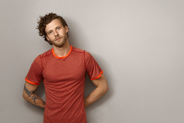 Positive bearded young male runner with curly hairdo and tattoo on muscular arm leaning on blank...