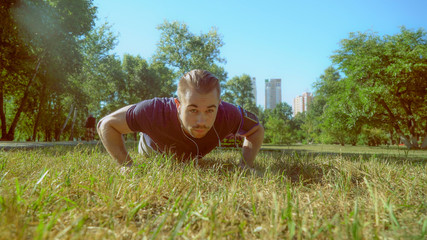 A handsome man in trendy outfit makes push ups during the video. He is wearing a blue T-shirt and an armband for the smartphone. The male is working out in the park with an urban background.