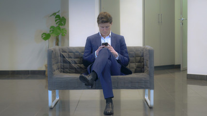 Fototapeta premium Young fashionable businessman using smartphone texting message sitting on the sofa. Caucasian young man play game on mobile phone going out leave waiting room.