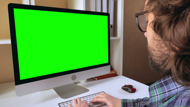 businessman sitting at workplace in small office. man with beard looking on display computer with green screen. employee in startup company typing on keyboard - Powered by Adobe