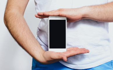 Phone in hand with a blank screen. A man wearing a white T-shirt is holding a phone with a blank display. Content completion concept.