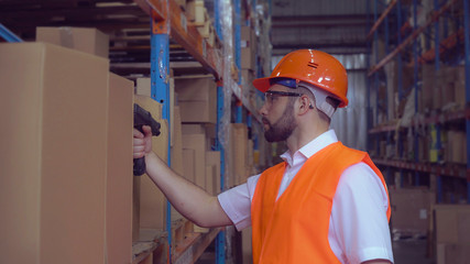 manager using wireless barcode scanner scanning labels on boxes before delivery in logistic center. Handsome professional worker wearing uniform white shirt high visibility orange hard hat and vest.
