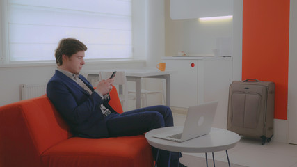 tourist sitting in the hotel room using internet for messaging on smartphone. Handsome man sitting in modern contemporary flat messaging on smart phone. Businessman wearing in elegant suit chatting on
