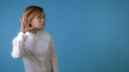 close up face young asian businesswoman posing showing hand gesture listening you on blue background in studio. attractive korean woman with blond hair wearing white casual shirt looking at the camera