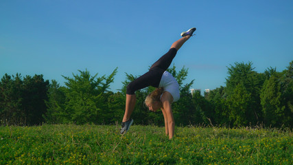Fototapeta na wymiar Blonde woman doing exercises for flexible and coordination in park with beautiful fresh green grass and summer nature. gymnast trains skills in gymnastics.
