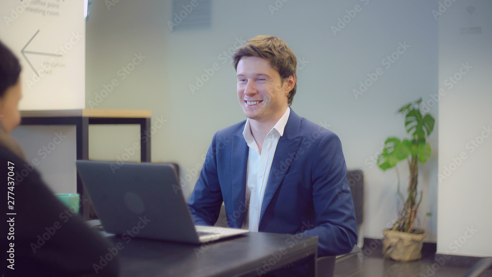 Poster friendly interviewer asking question to woman. Handsome smiling talking with candidate for work. People sitting at the desk in modern room opposite each other.