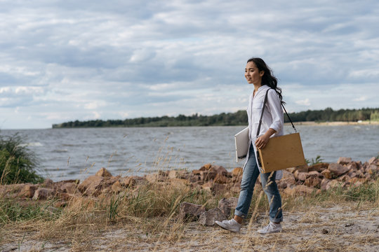 Asian woman artist looking for inspiration. Beautiful smiling Korean girl holding canvas, paint brush, easel, enjoying beautiful landscape. Portrait of happy successful painter walking along beach