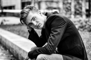 Serious or sad blond young man in jeans and jacket, sitting outdoors looking in camera