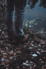 A man wearing light jeans and boots stands by the river. Kind of legs. Autumn. Evening. Close-up