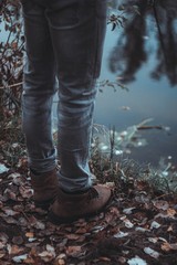 A man wearing light jeans and boots stands by the river. Kind of legs. Autumn. Evening. Close-up