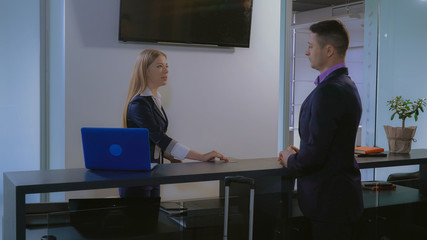 Adult man pays by credit card for room in hotel. Smiling woman using computer at working place. Worker and tourist standing near reception desk. Young receptionist talking with guest.