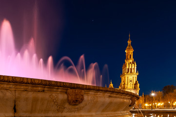 Fototapeta na wymiar Seville, Spain. Amazing view of Spanish square with detail of the fountain with water illuminated by light violet light. In the background illuminated tower and starry blue sky