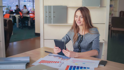 Business woman using app on the touch screen tablet. On her desk documets and charts with sales report