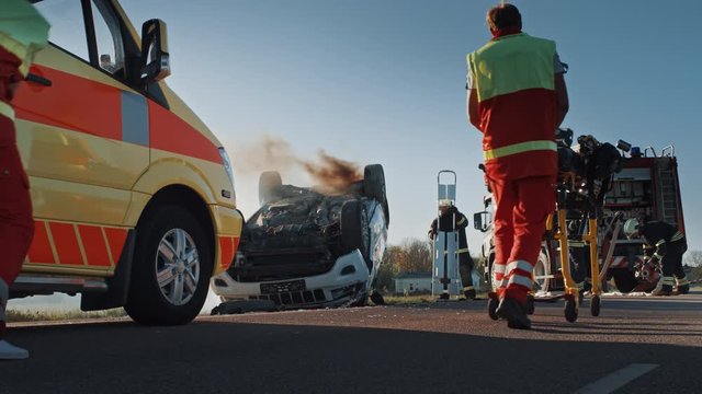 On the Car Crash Traffic Accident Scene: Team of Paramedics and Firefighters Rescue Injured People Trapped in Rollover Vehicle. Professionals Extricate Victims, give First Aid, Extinguish Fire