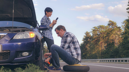 handsome caucasian man changing a tire on the side of the road. Woman using mobile phone messaging...