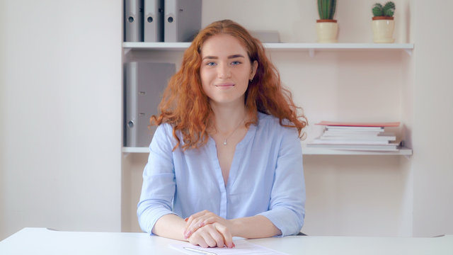 Portrait successful business woman with curly redheaded long hair sitting at the white desk in office sign document. Attractive female wearing casual elegant blue shirt undersign contract looking at