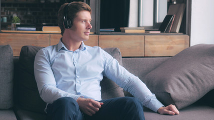 Young businessman relaxing at studio apartment. Handsome man listening music sitting on the sofa in flat. Resting after working day at home with modern interior.