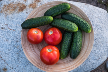 Fresh vegetables in a wooden round plate. Red tomatoes and green cucumbers in a bamboo bowl. Against the background of granite boulders, stones. Summer evening