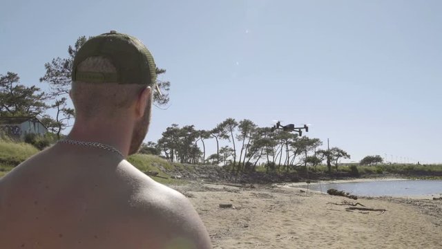 Handsome young tattooed bearded blond man shirtless with a green cap and denim shorts doing drone on the beach in sunny summer. 4K UHD. Back view.