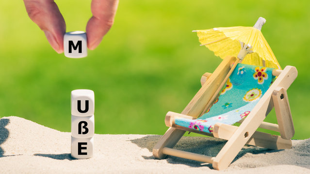 Am Sandstrand bilden W&uuml;rfeln das Wort "Mu&szlig;e" neben einer kleinen Sonnen-liege und einem Sonnenschirm.