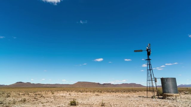 Loop of A static daytime timelapse of a typical arid Karoo farm landscape, a windmill blowing in the wind next to an old metal dam, scattered cumulous clouds forming against a bright blue sky
