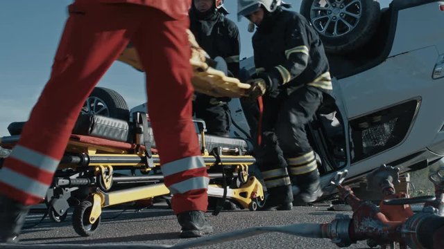 On the Car Crash Traffic Accident Scene: Team of Firefighters Pull Female Victim out of Rollover Vehicle, They Use Stretchers Carefully, Hand Her Over to Paramedics who Perform First Aid. Low Angle
