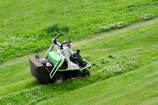 Ride-on Lawn Mover. Rear Back View Gardener Man Driving A Riding Lawn Mower In Garden. Gardener Cutting The Grass Of A Garden Seated On A Lawn Mower.