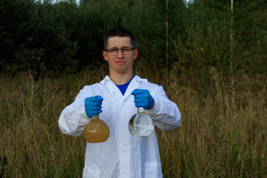 Laboratory technician holding 2 glass flasks with analyses of clean and dirty water. The man is wearing blue latex gloves and a white robe. Analysis of water in natural reservoirs in nature