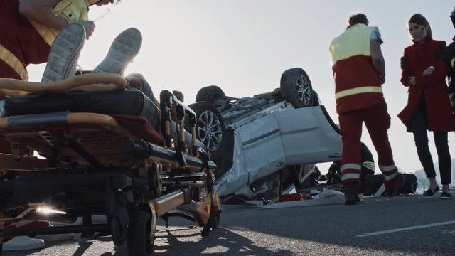 Car Crash Traffic Accident: Firefighter Leads Mother to a Child Lying on Stretchers Whom Paramedics Trying to Save / Resuscitate. Rollover Vehicle with Other Trapped Passengers in the Background