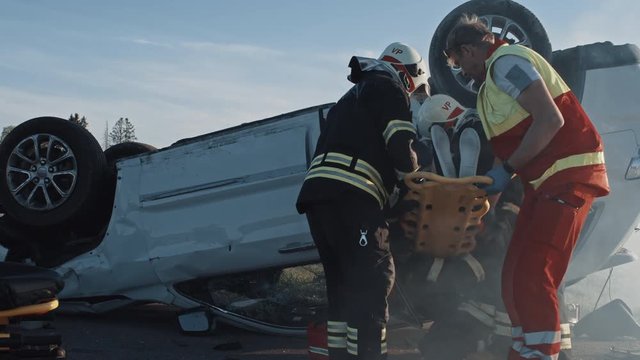 On the Car Crash Traffic Accident Scene: Rescue Team of Firefighters Pull Female Victim out of Rollover Vehicle, They Use Stretchers Carefully, Hand Her Over to Paramedics who Perform First Aid