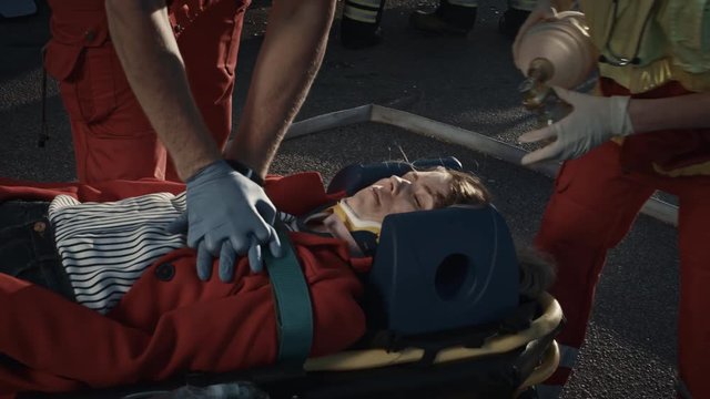 On the Car Crash Traffic Accident Scene: Paramedics Saving Life of a Female Victim who is Lying on Stretchers. They Apply Oxygen Mask, Do Cardiopulmonary Resuscitation / CPR and Perform First Aid