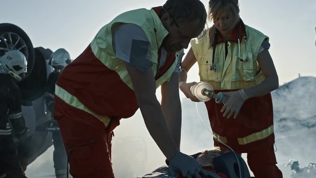 On the Car Crash Traffic Accident Scene: Paramedics Saving Life of a Female Victim who is Lying on Stretchers. They Apply Oxygen Mask, Do Cardiopulmonary Resuscitation / CPR and Perform First Aid