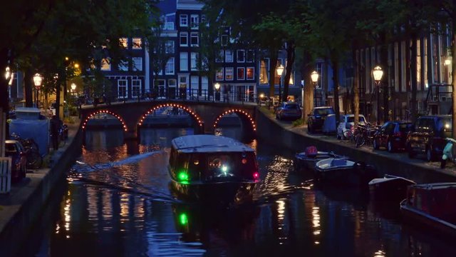 Amazing city nightlife. Sightseeing boats floating the canal. People walking on the bridge and riding bicycles. Typical dutch houses, bicycles. Flemish architecture of historical district, Amsterdam