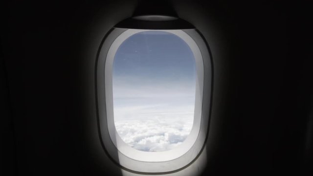 Slow motion dolly in to an airplane window from inside with clear blue sky with white clouds and a wing and engine view