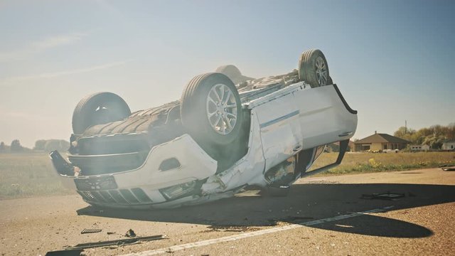 Horrific Traffic Accident, Rollover Smoking and Burning Vehicle Lying on its Roof in the Middle of the Road after Collision. Daytime Crash Scene with Severely Damaged Car.