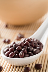 Raw, uncooked, dried adzuki (red mung) beans in white spoon on bamboo mat background