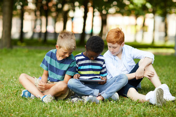 Naklejka na ściany i meble Three smiling teenage school friends sitting in park on grass and playing new tablet game together
