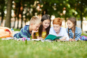 Four teenage school friends reading together story for Literature class in park lying on grass...