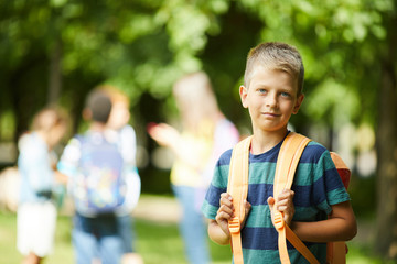Smiling teenage schoolboy with orange backpack standing outside in park before beginning of classes