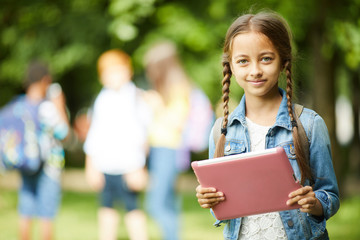 Smiling teenage schoolgirl with braided hair and pink tablet standing outside in park before...