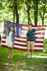 Portrait of smiling friendly American kids in casual outfits standing in forest and holding national flag